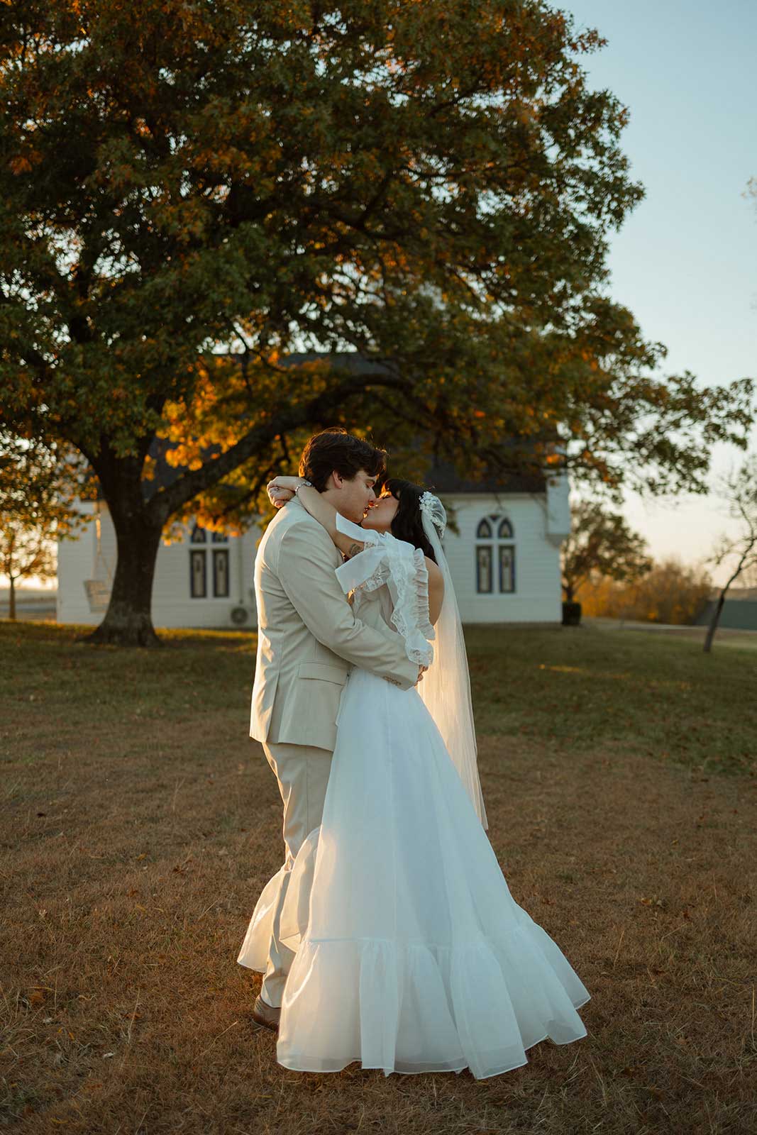 Newlyweds kissing beneath a large oak tree outside the Willowood Ranch chapel at golden hour.